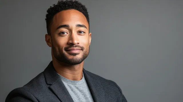 Professional headshot of a man with his shoulders rotated roughly 20 degrees off-camera and his head turned back to the lens, dark blazer over a grey tee on a grey studio background