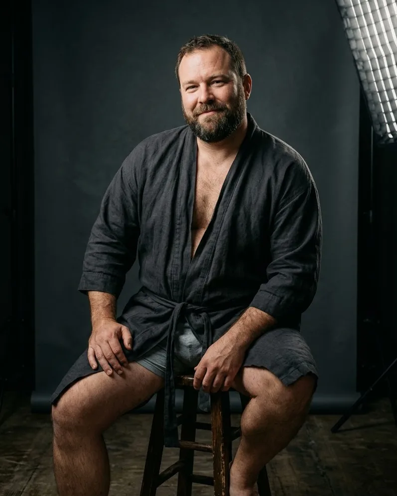 Bearded man in a charcoal robe seated on a wooden stool — male boudoir studio session